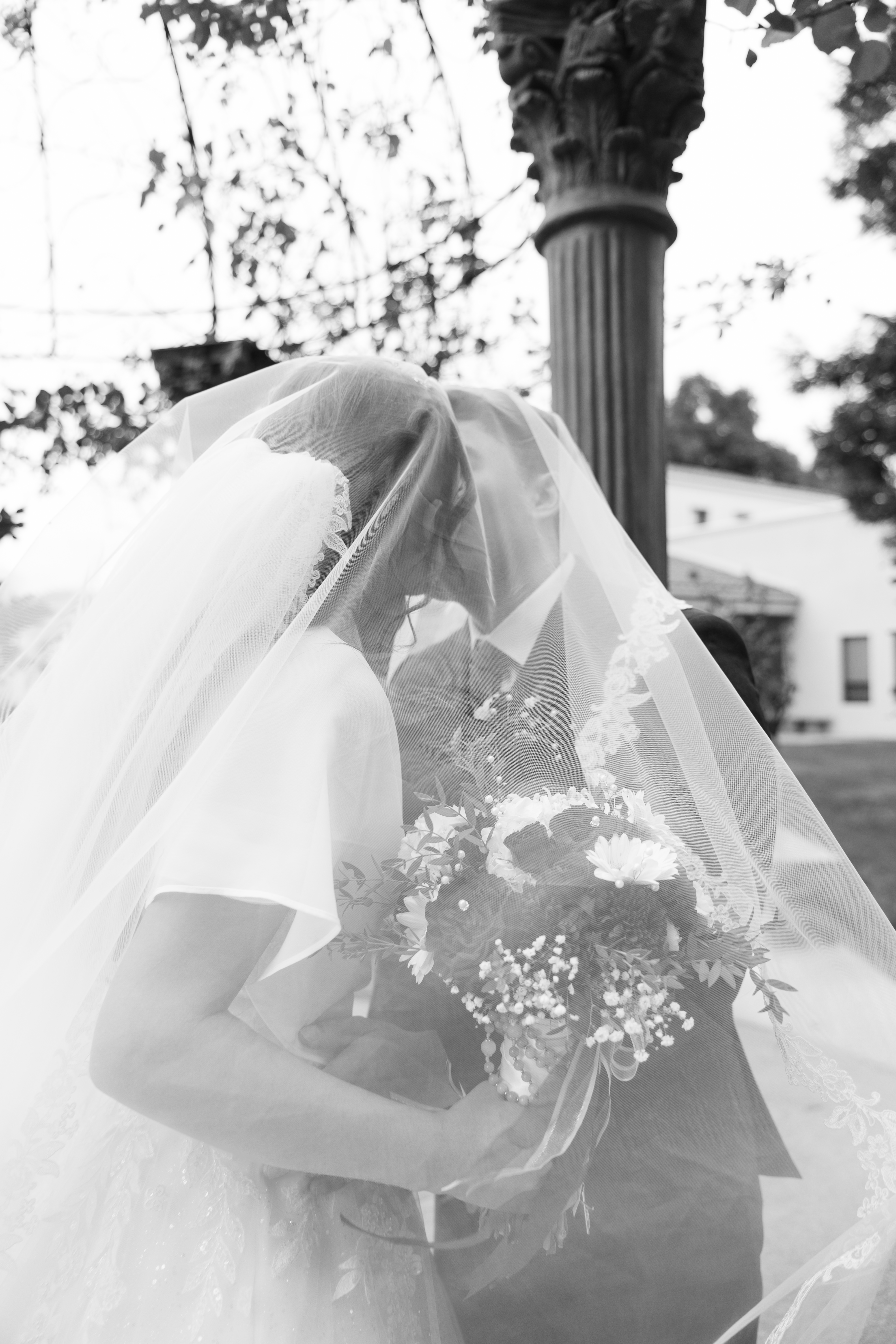 Bride and groom embracing under veil with bouquet, black and white